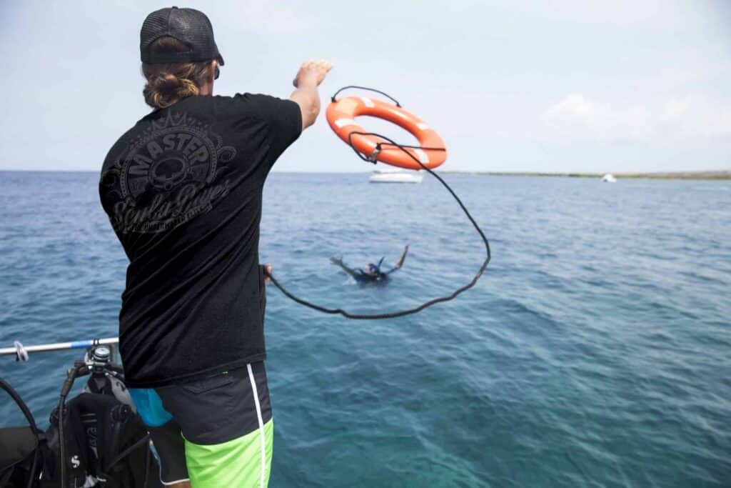 Back view of a person on a boat casting a fishing line into the ocean, with an orange life ring nearby and divers in the water behind them.