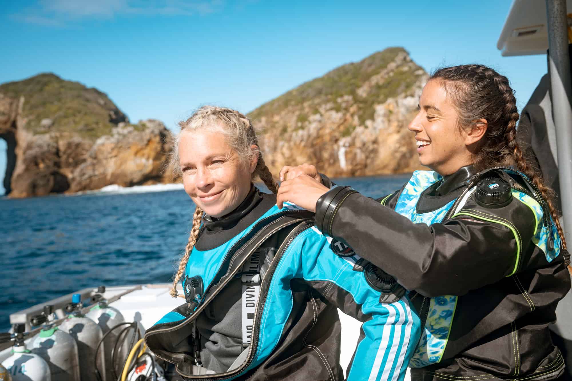 A diver helping her buddy zip into a drysuit on the boat